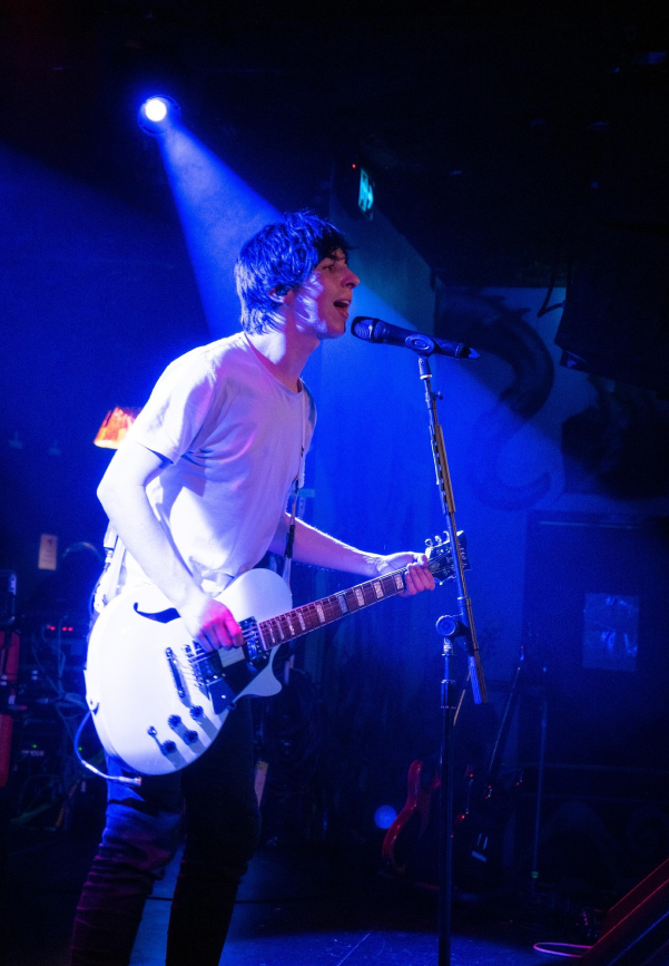 A man in a white t shirt playing electric guitar and singing into a microphone under a blue spotlight