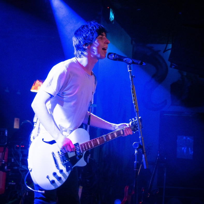 A man in a white t shirt playing electric guitar and singing into microphone on stage under a blue spotlight