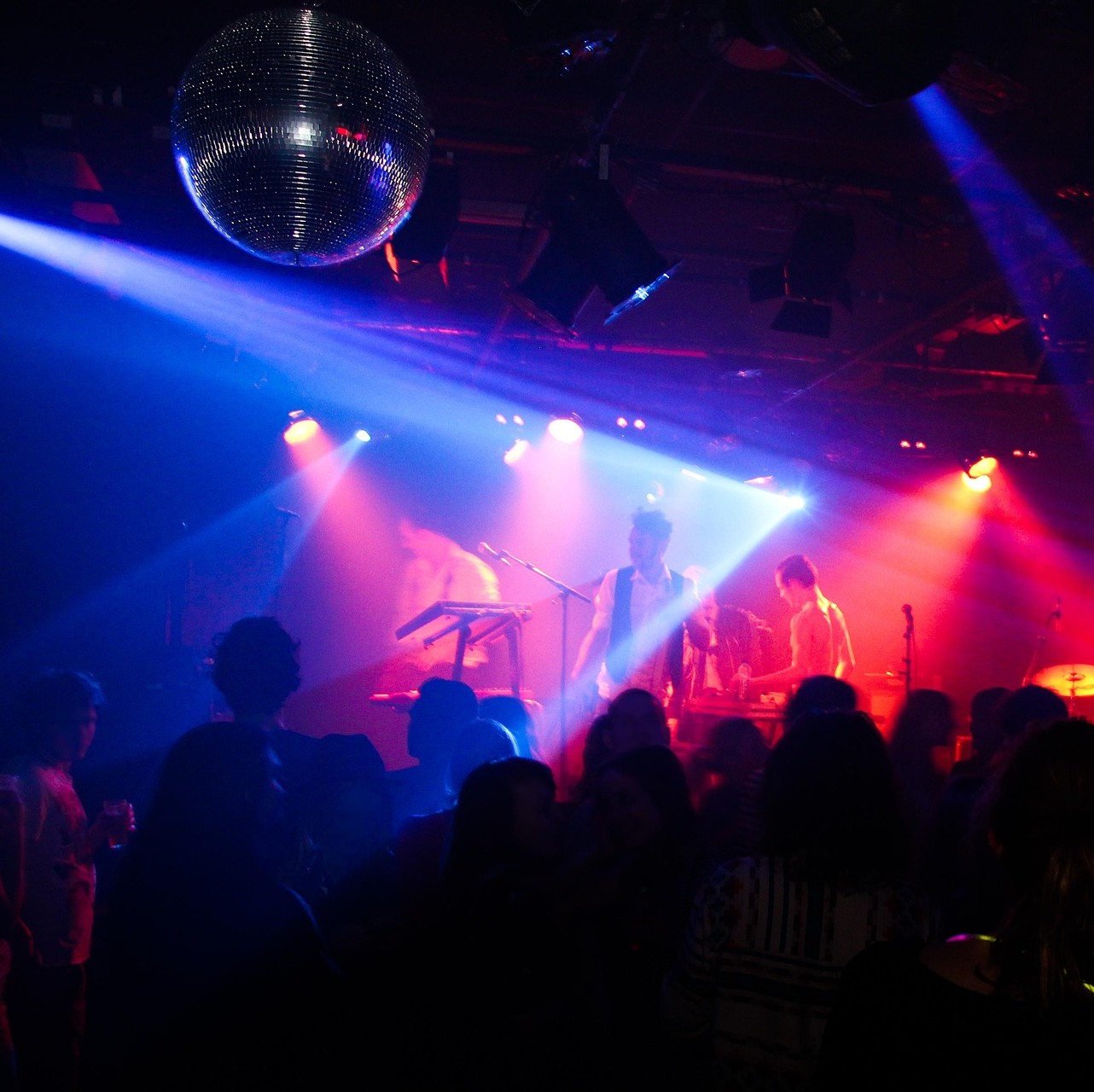 An image of a three person band on stage with a crowd in front and a discoball on the ceiling