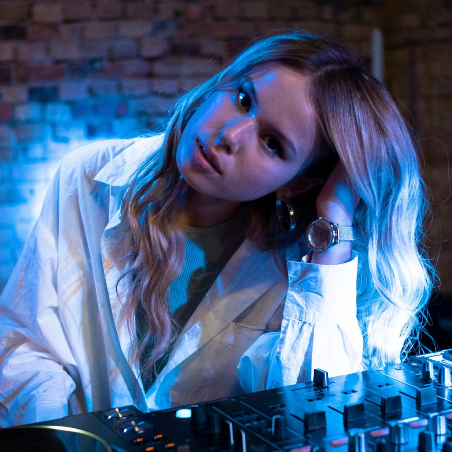 A blonde woman in a white button up in front of a dj deck with headphones around her neck