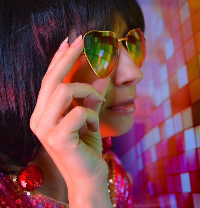A lady with black hair wearing heart shaped sunglasses and looking at a colourful discoball